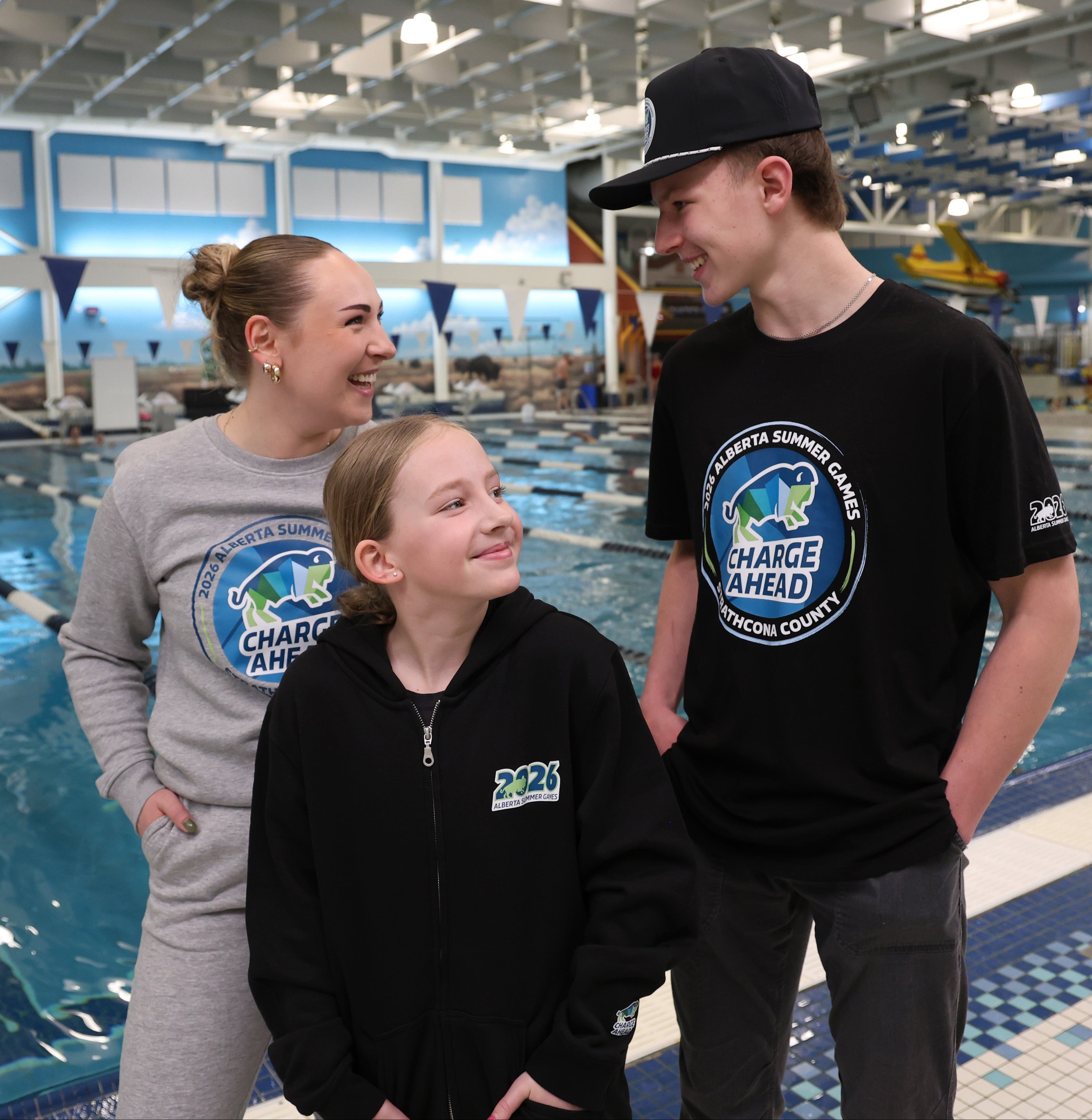 Three people standing by a pool with 'Charge Ahead' swag in an indoor swimming facility.